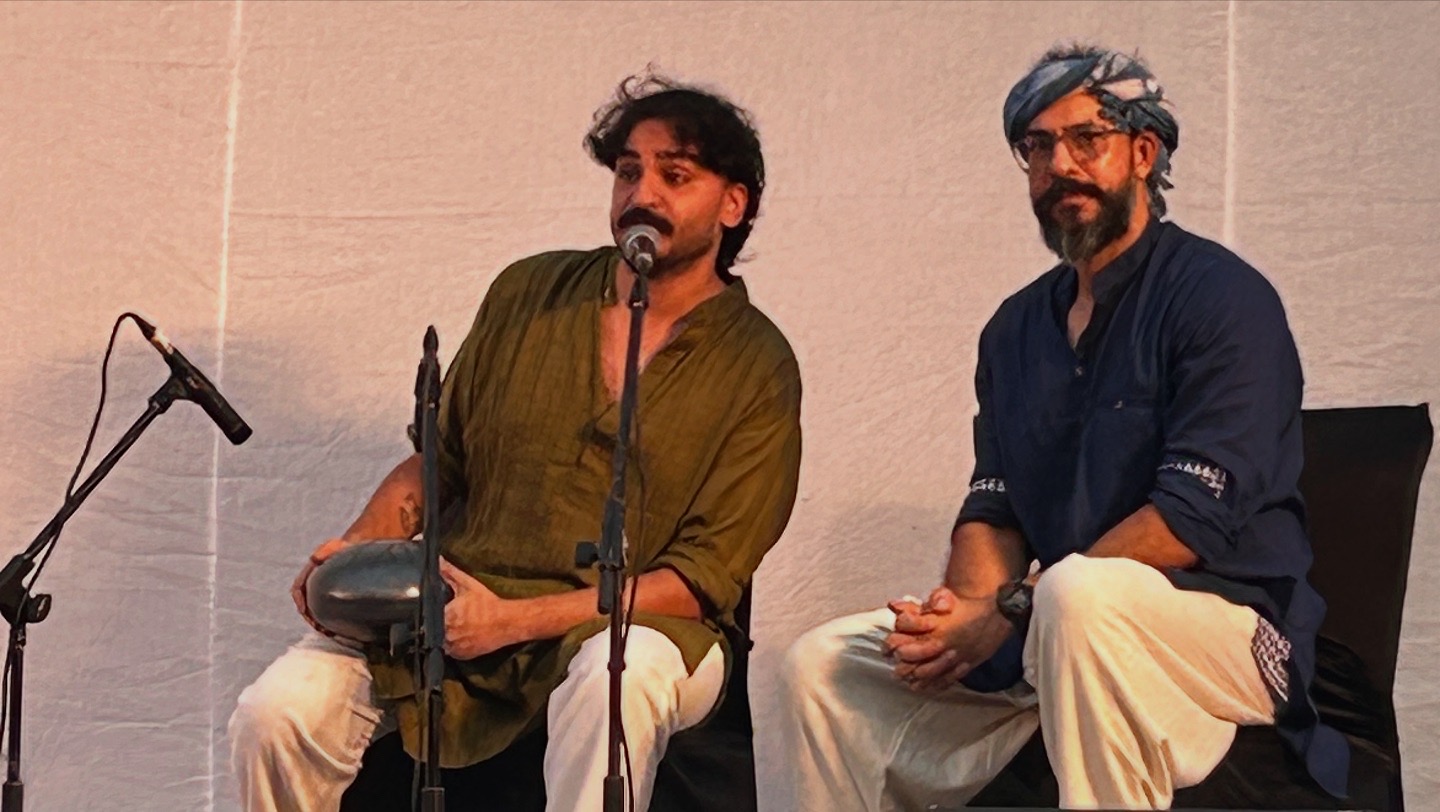 Ashim Bery speaking into a microphone on stage at Baarish, his handpan on his lap, beside a fellow performer against a white drape
