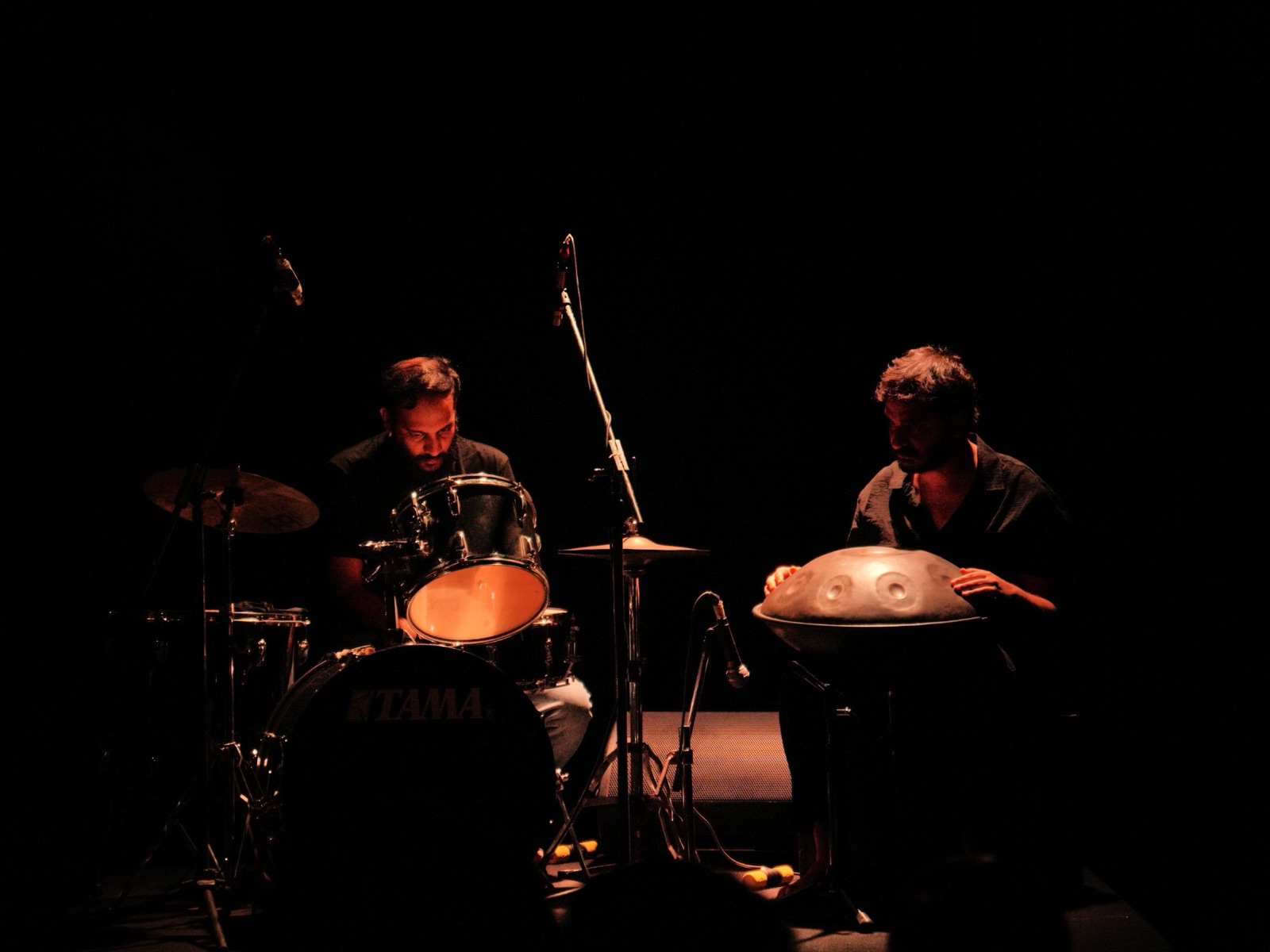 Ashim Bery at a handpan, lit warm on a dark stage, with a drummer at a kit beside him