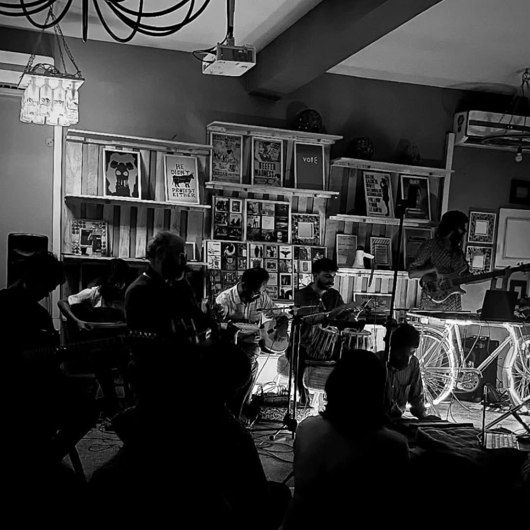 Black-and-white photo of the Delhi Improvisers Orchestra performing in a small room lined with posters, musicians on guitar, tabla and bass, the audience in silhouette