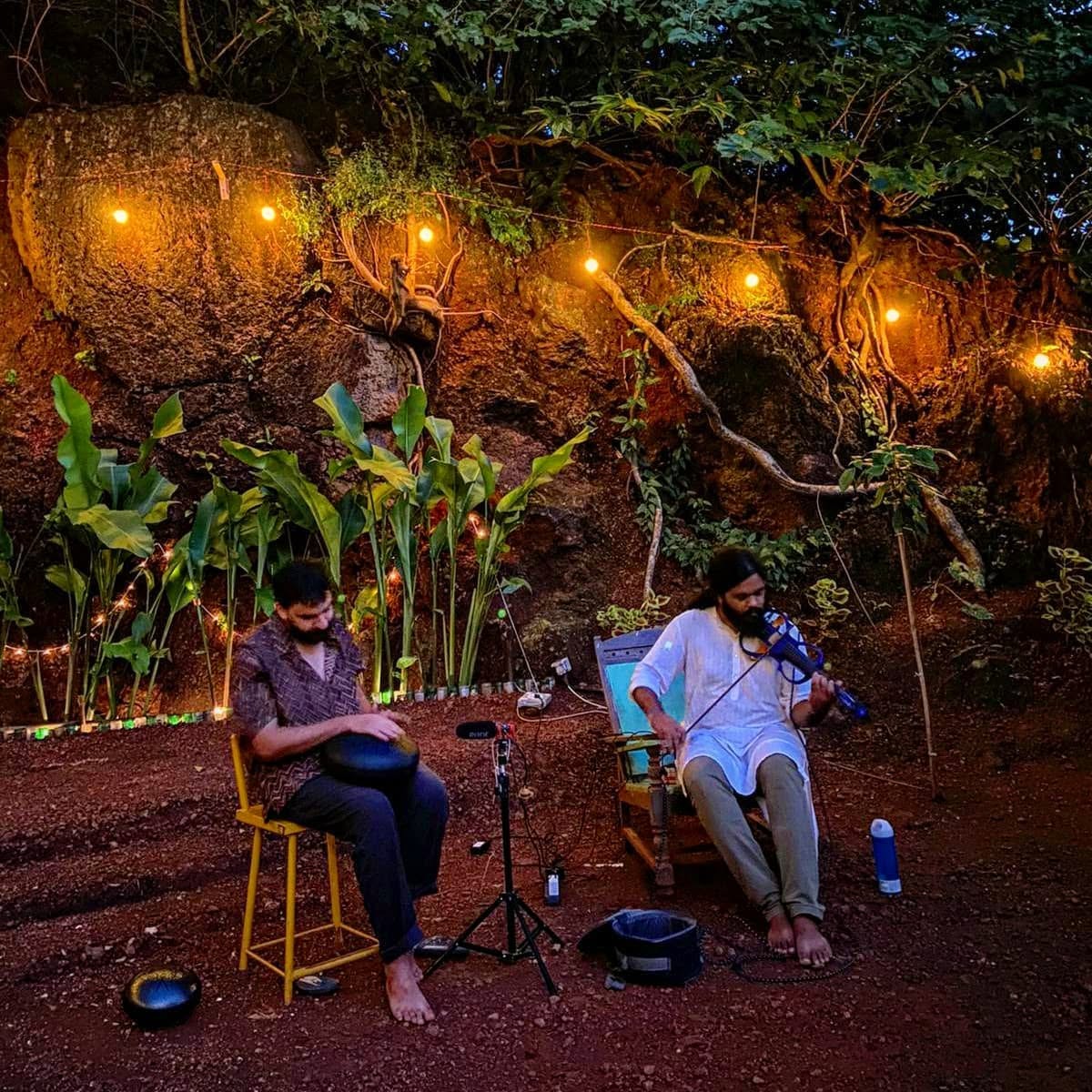 Ashim Bery playing a handpan outdoors at dusk beside a violinist, under string lights in front of a rock face with plants
