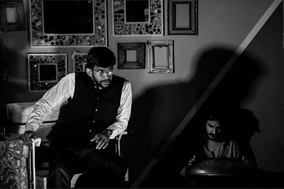 Black-and-white scene from The Patriarch: an actor rises from his chair as Ashim Bery plays a handpan in the shadows