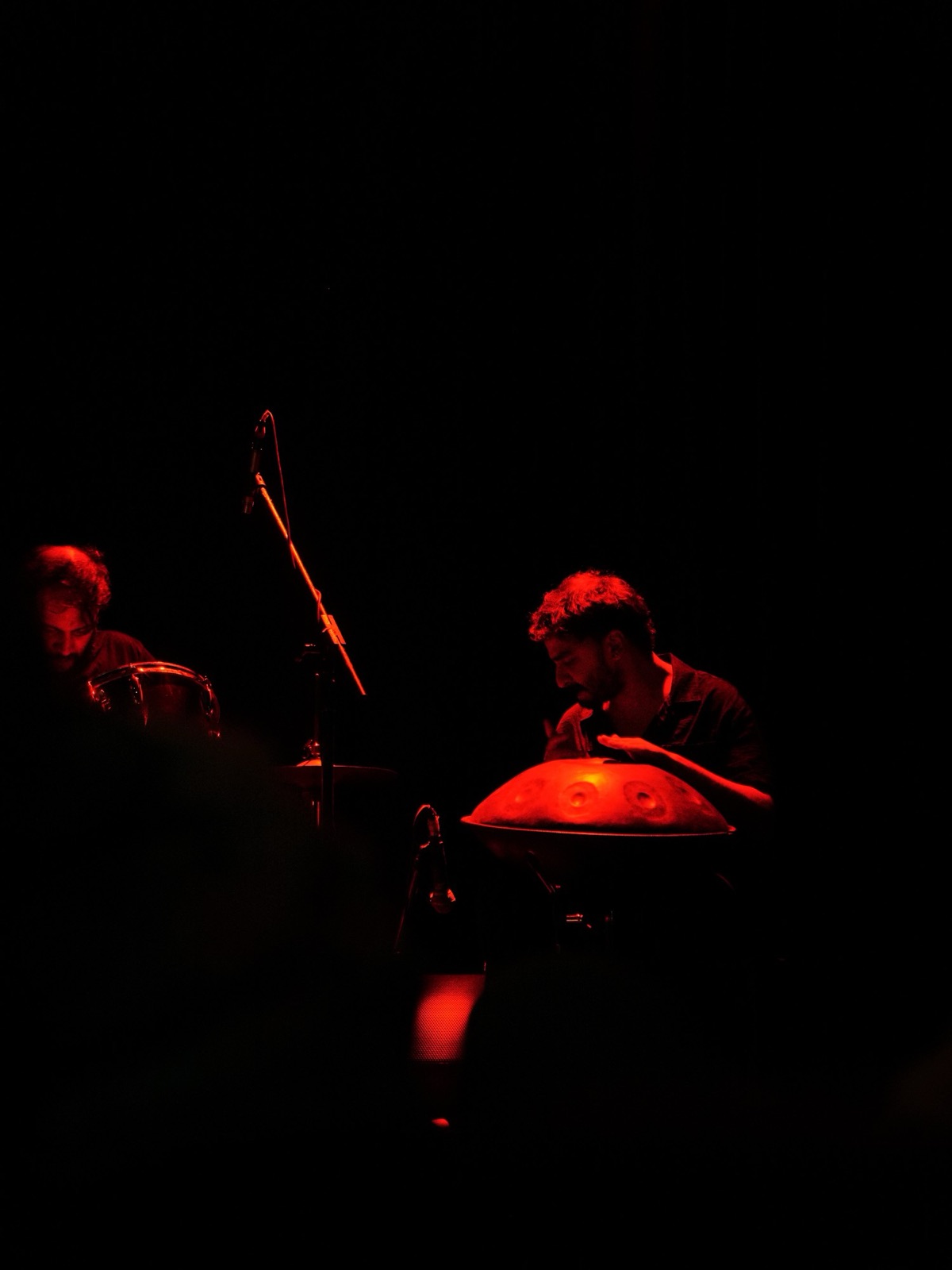 Ashim Bery playing a handpan in deep red light, a percussionist at the edge of the frame